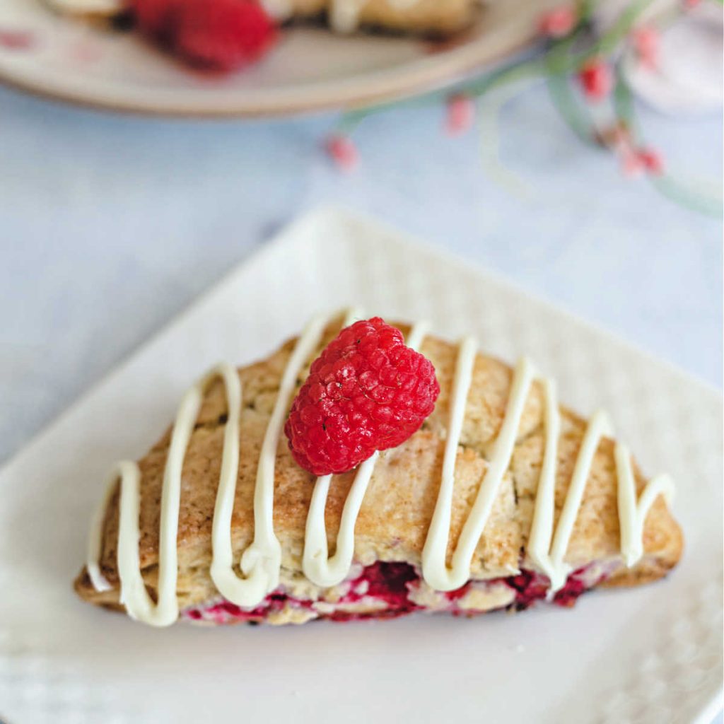 a raspberry scone with white chocolate drizzle on a white plate on a table