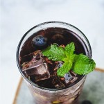 a glass of blueberry tea with ice cubes and a mint sprig garnish sitting on a coaster.