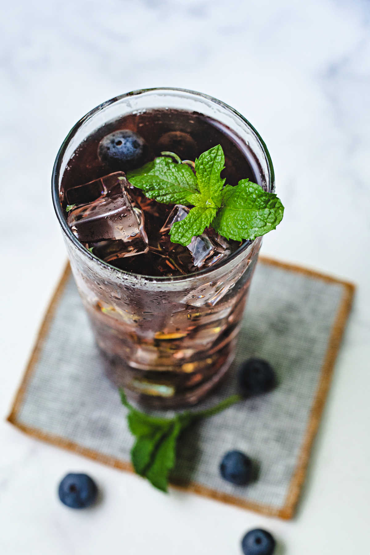 a glass of blueberry tea with ice cubes and a mint sprig garnish sitting on a coaster.