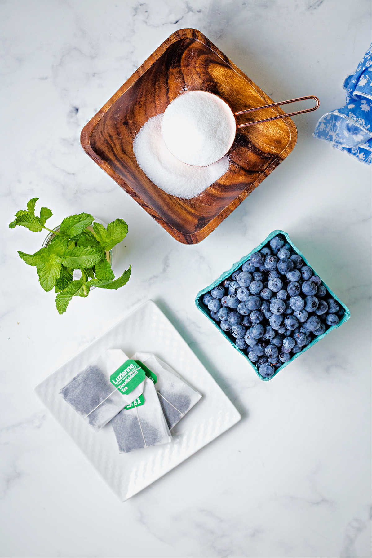 ingredients for blueberry tea on a table: blueberries, sugar, tea bags, and mint sprigs.