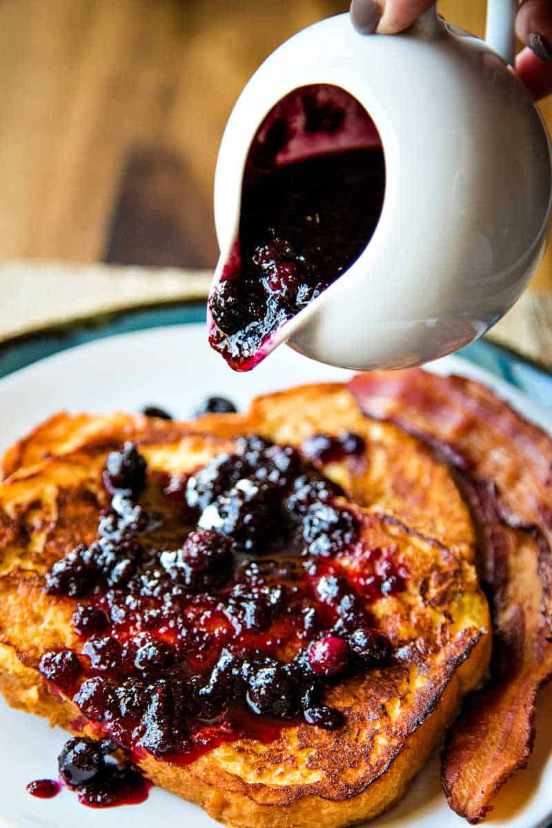 pouring warm berry syrup out of a white pitcher onto french toast