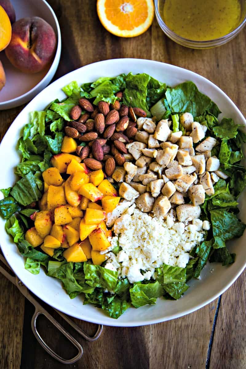 white bowl on a wood plank table with ingredients for chopped chicken and peach salad