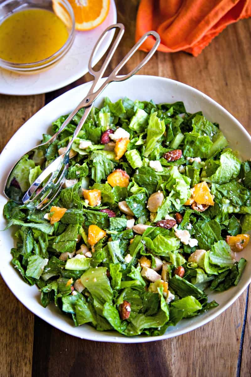 white serving bowl of romaine lettuce, chicken, peaches with salad tongs on a wooden surface