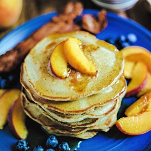 a stack of pancakes on a blue cake with peaches and blueberries scattered around and syrup drizzled on top.