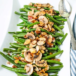 green bean almondine on a white serving platter with a silver serving spoon and with almonds and mushrooms scattered on top.