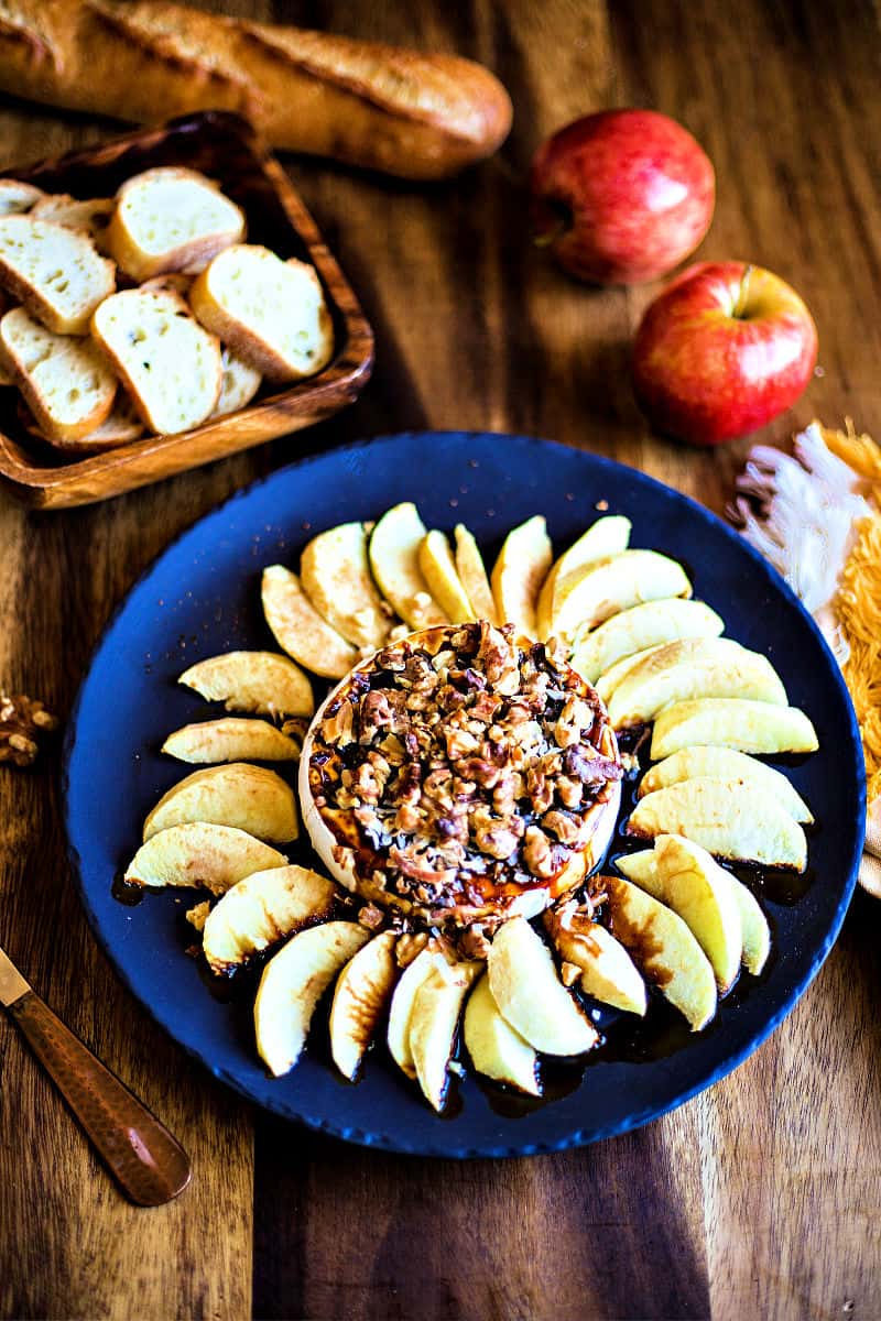 black slate platter with baked Brie and apples on a wooden table with two apples and a baguette