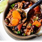 a spoonful of black bean sweet potato chili lifting out of a crock sitting on a white plate with a lime wedge.