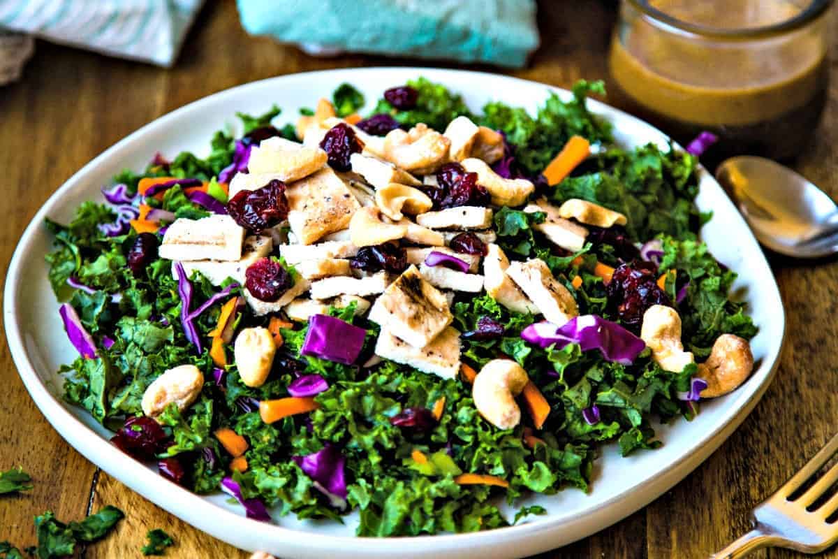 a table set with a plate of chopped chicken kale salad, a fork, scattered cashews, and a bowl of vinaigrette