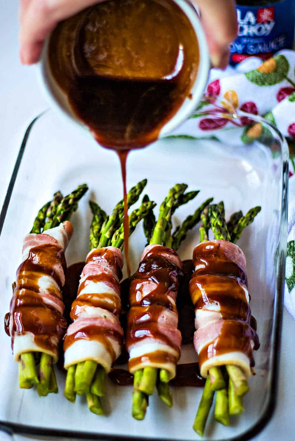 pouring brown sugar soy sauce over bacon wrapped asparagus in a baking dish