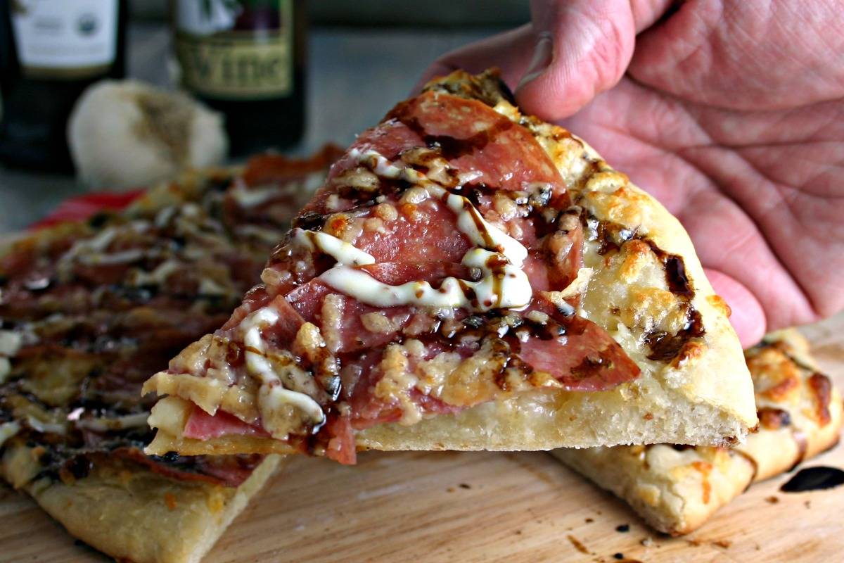 A close up of a person holding a slice of Salami Pizza with Garlic Dijon Aioli
