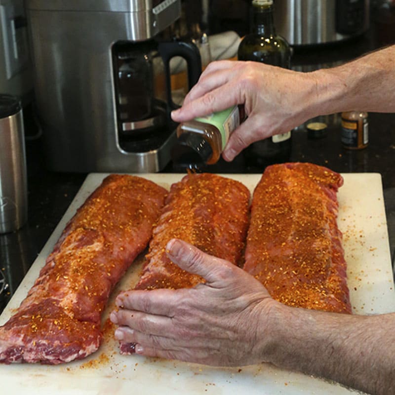 Shaking spice rub on racks of baby ribs on a counter