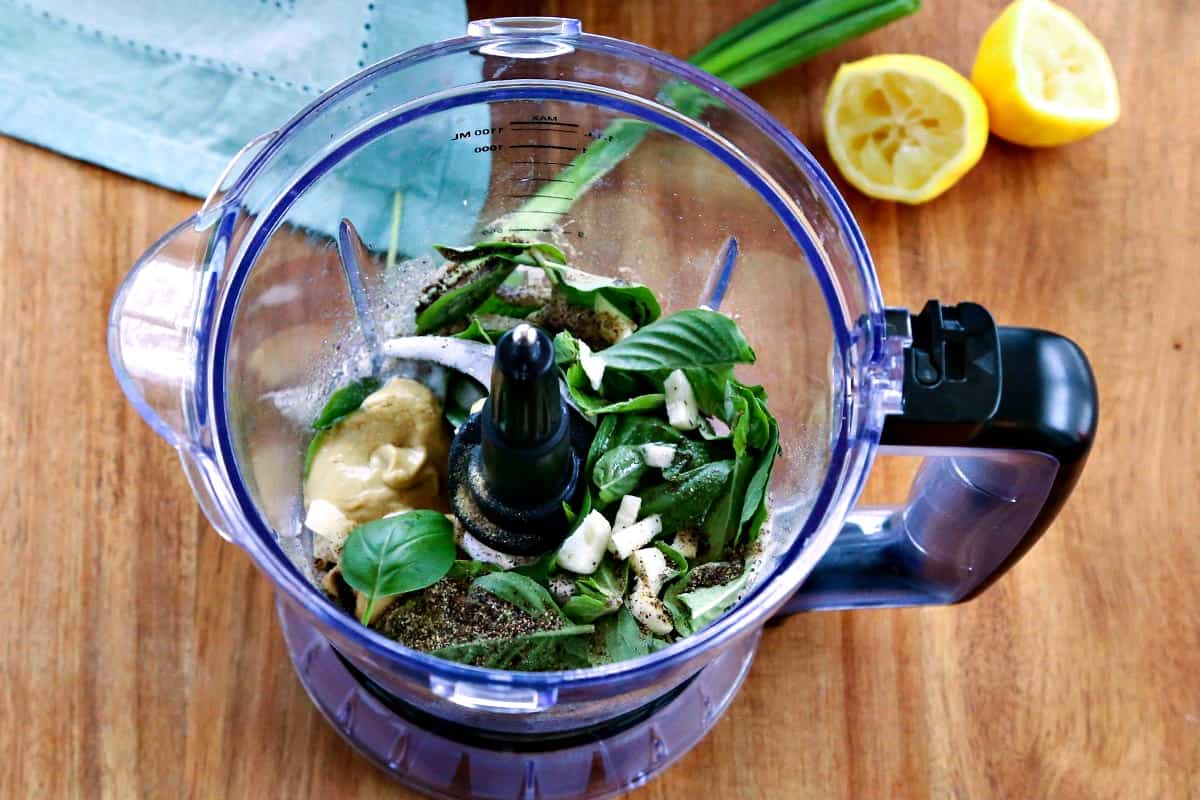 A bowl of a food processor sitting on top of a wooden table, with Basil and garlic