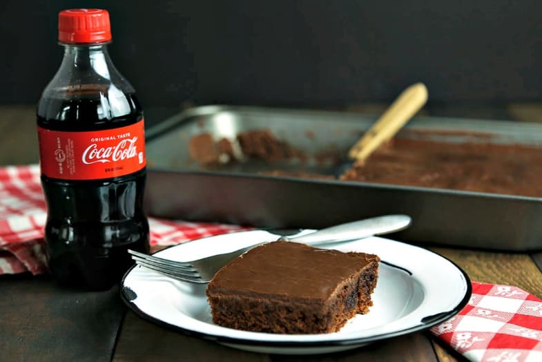 A bottle of Coca Cola with a slice of cake on a plate, with Classic Coca-Cola Chocolate Cake