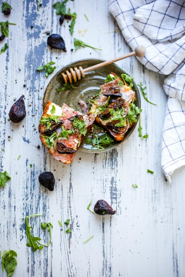 A plate of food with a slice of toast, with Ricotta cheese, figs, and arugula on top of a wooden table