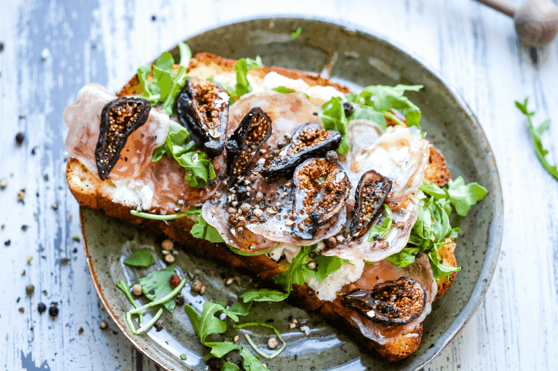 A plate of food with a slice of toast, with Ricotta cheese, figs, and arugula 