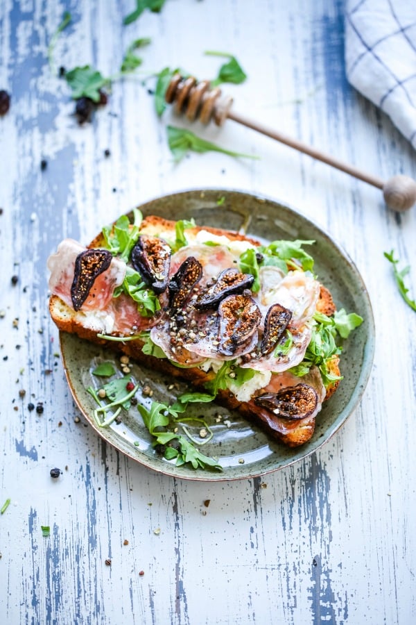A plate of food with a slice of toast, with Ricotta cheese, figs, and arugula on top of a wooden table