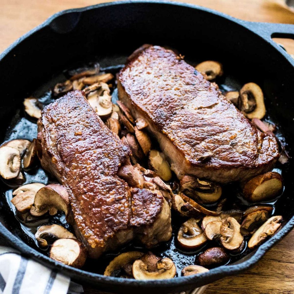 two New York strip steaks with mushrooms in a cast iron skillet
