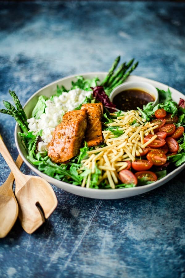 a white bowl with salad ingredients and wooden salad tongs