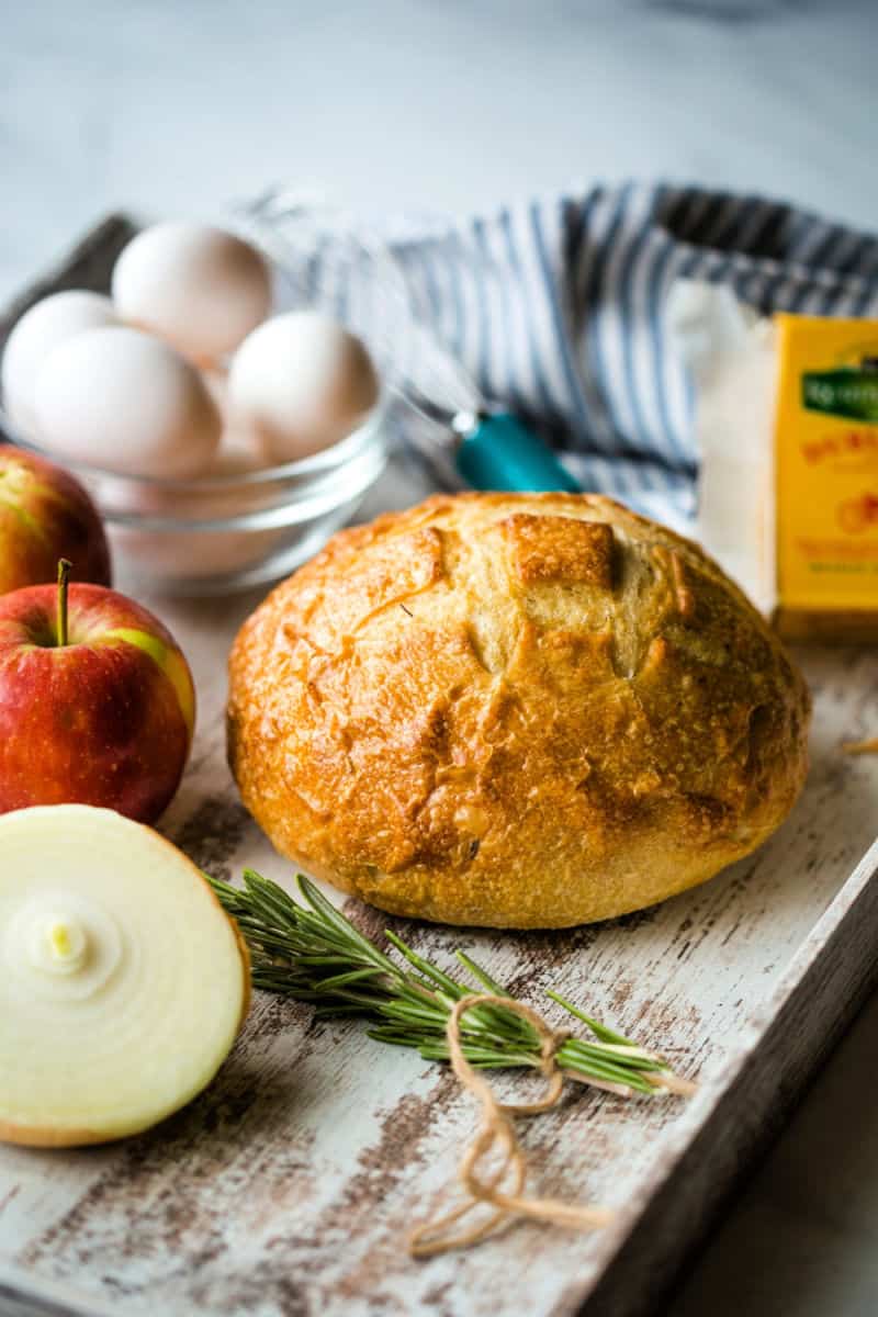 a loaf of sourdough bread on a tray with fresh rosemary, a bowl of eggs, apples, and a block of cheese