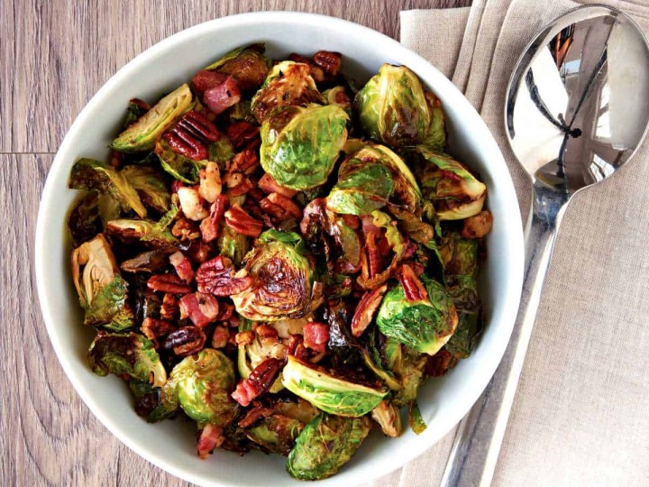 A bowl of Brussels sprouts wth pancetta and pecans on a wooden background with a serving spoon