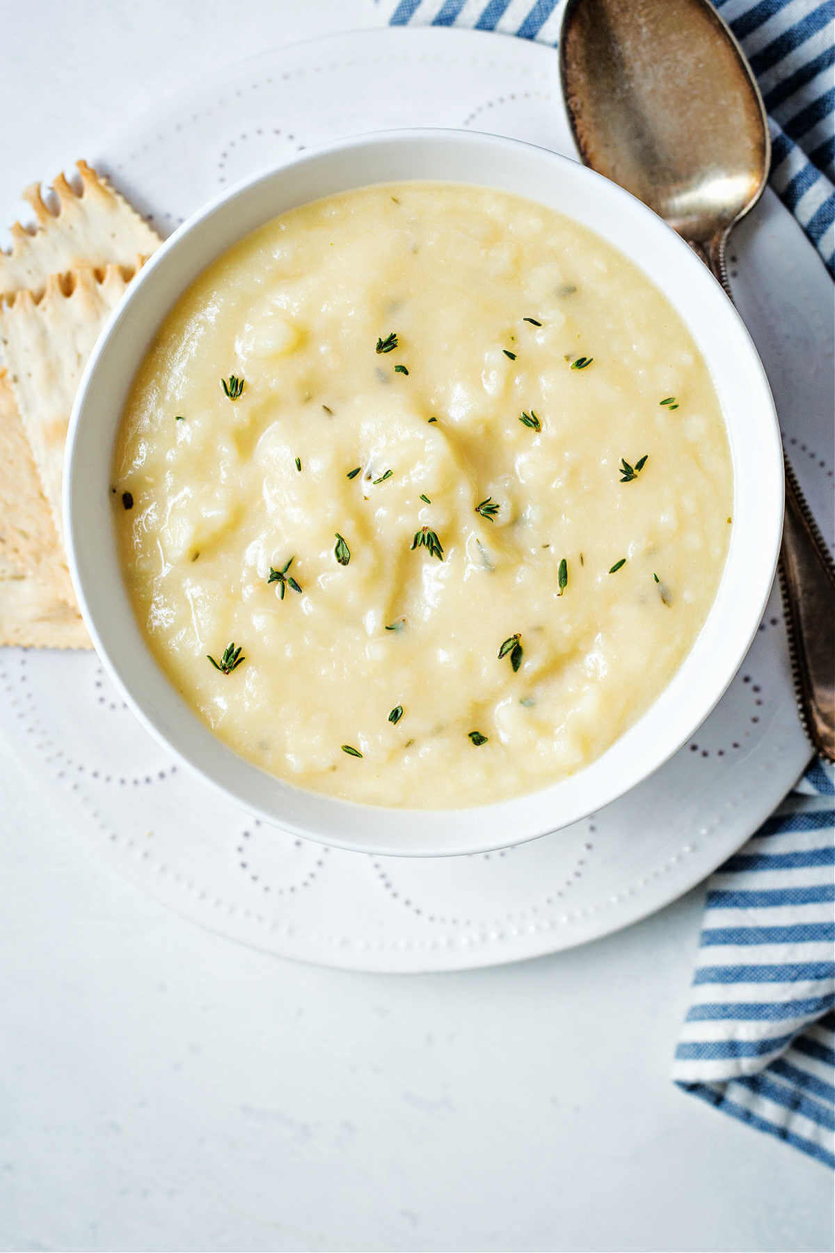 a bowl of potato soup on a white plate with crackers and a spoon to the side and fresh thyme leaves sprinkled on top.