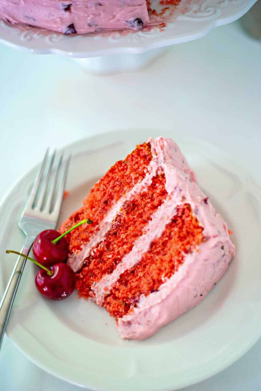 a slice of fresh cherry cake with fresh mint and buttercream frosting on a white plate with a fork and fresh cherry garnish