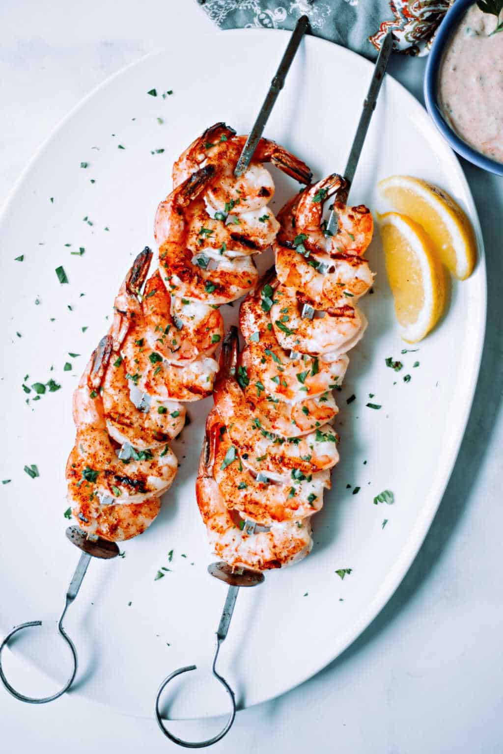 shrimp on the barbie skewers on a white platter with lemon wedges and parsley