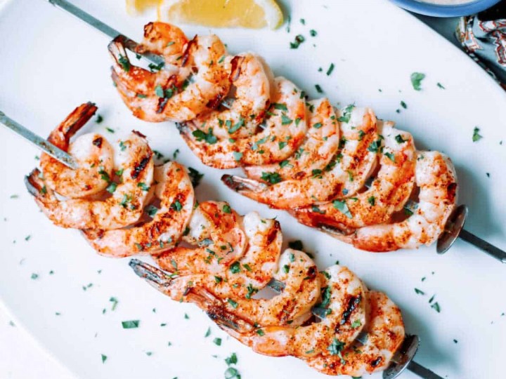shrimp on the barbie skewers on a white platter with lemon wedges and parsley