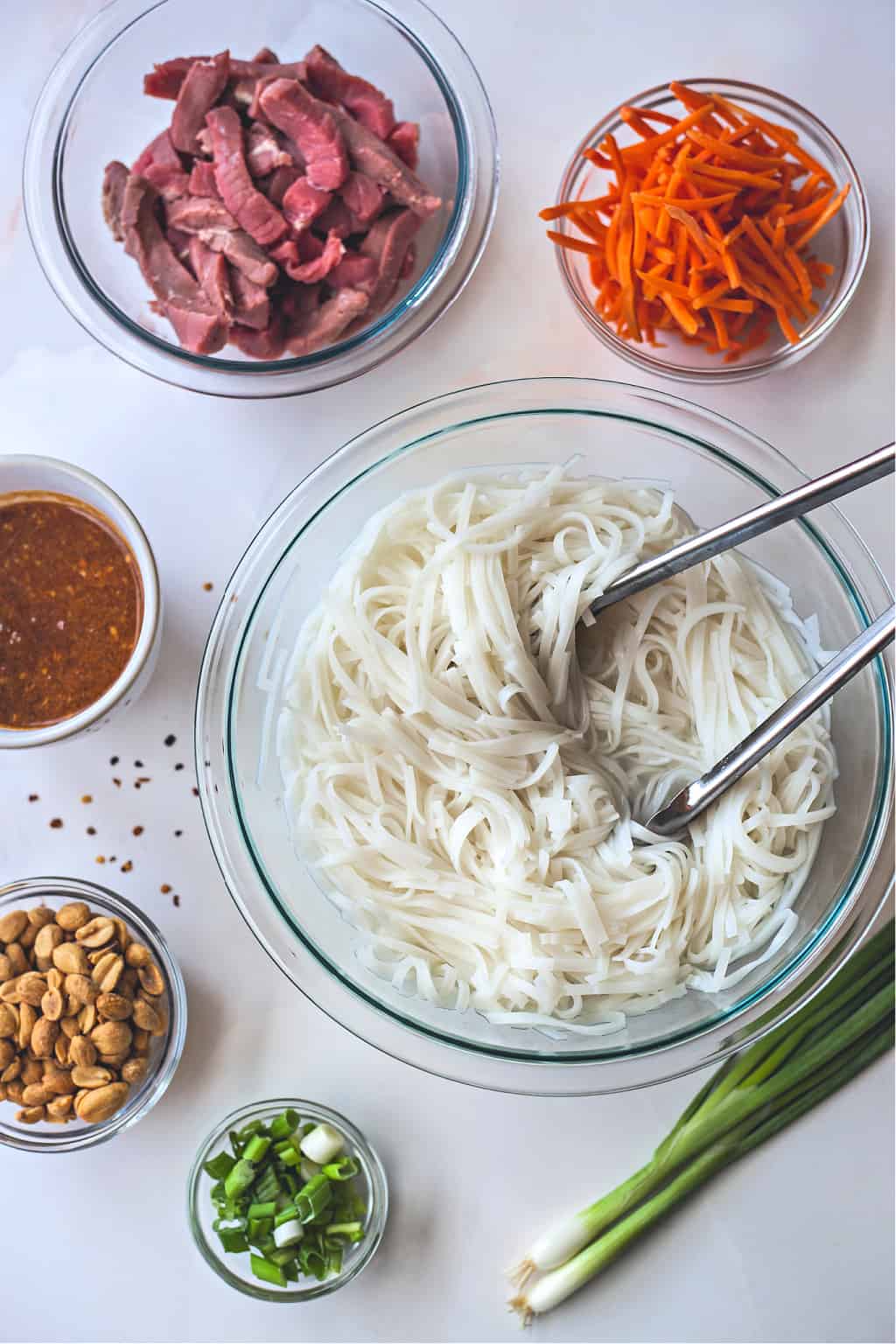 Bowls of pad thai ingredients on a table, with noodles, beef, peanuts