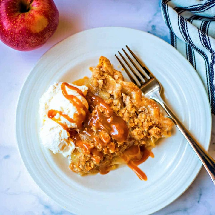 a slice of apple crumble pie on a white plate with a fork and apple to the side