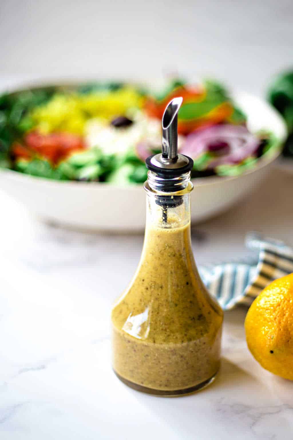 greek salad dressing with a bowl of salad in the background