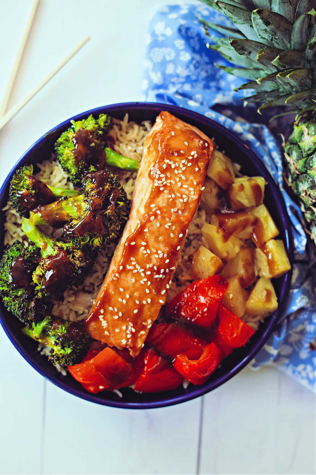 teriyaki salmon in a bowl with brown rice, roasted broccoli, red bell pepper, and pineapple on a wooden table