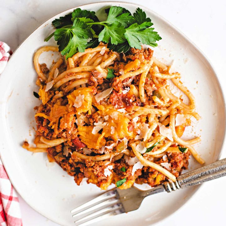 baked spaghetti on a white plate with a fork and red checkered napkin on a table.