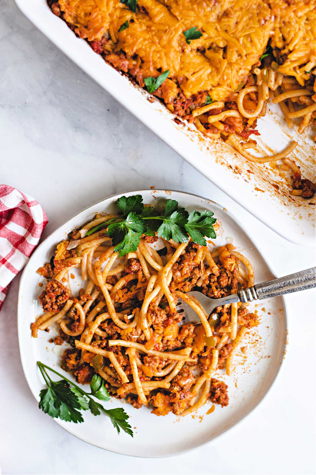 baked spaghetti on a white plate with a fork and red checkered napkin on a table.