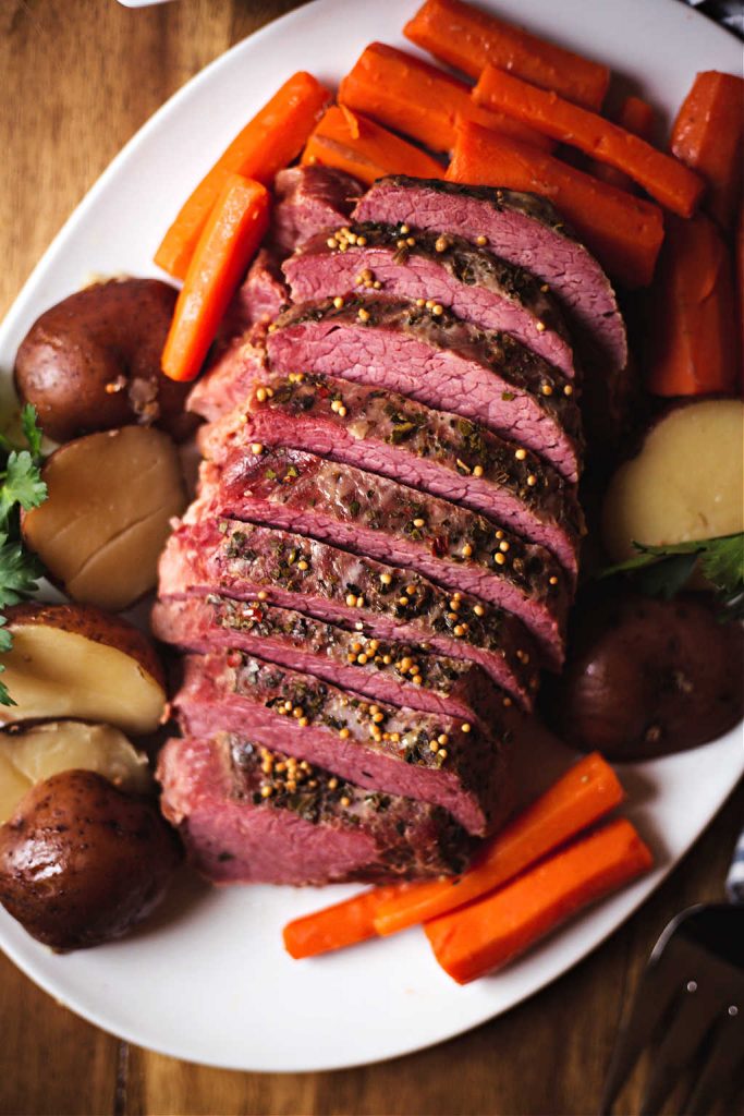 sliced slow cooker corned beef on a white platter with carrots and potatoes with a bowl of cabbage on a table.