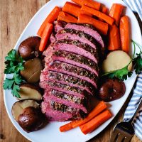 sliced slow cooker corned beef on a white platter with carrots and potatoes on a table.