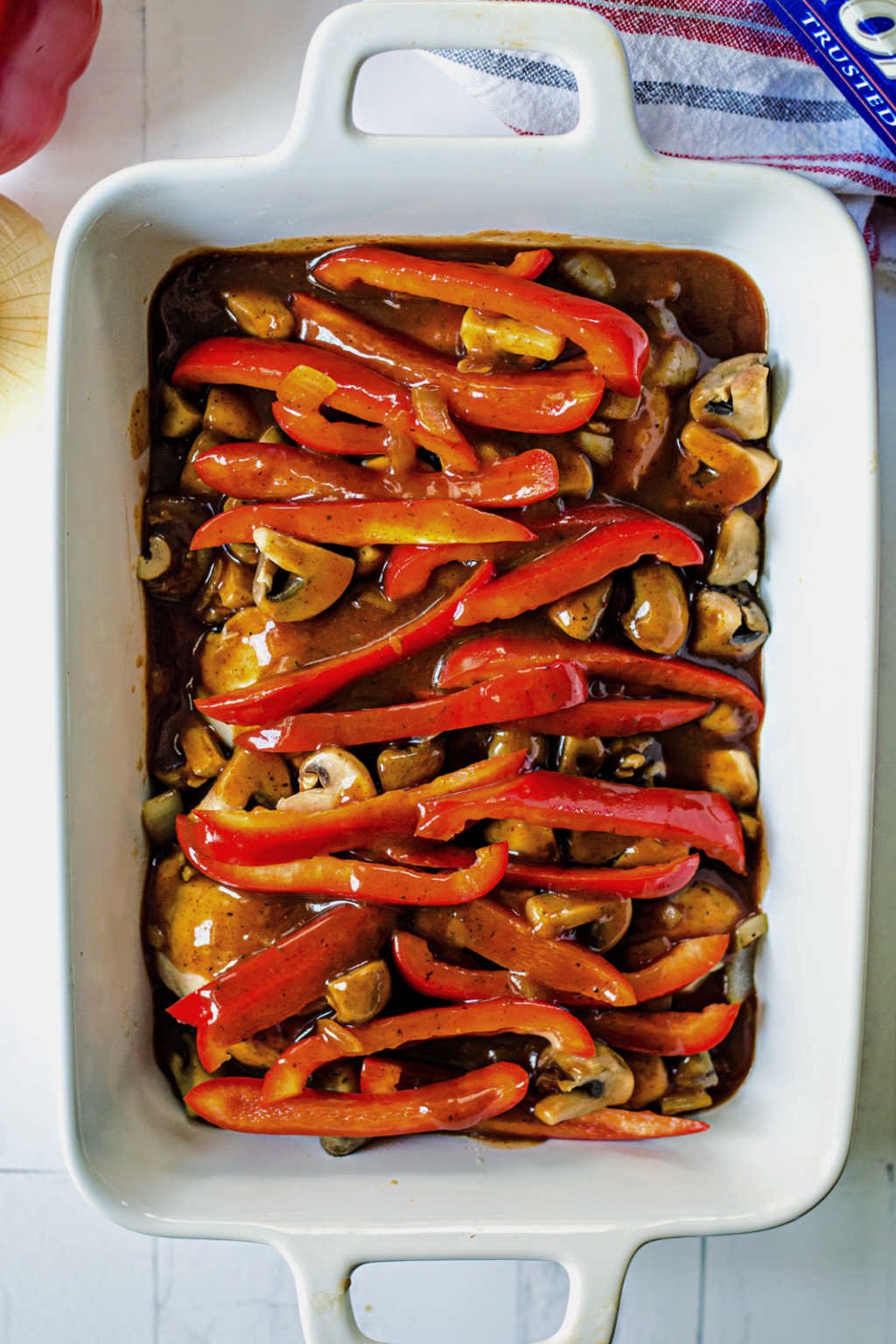 chicken breast with red bell peppers in a white baking dish on a table with sauce ready for the oven