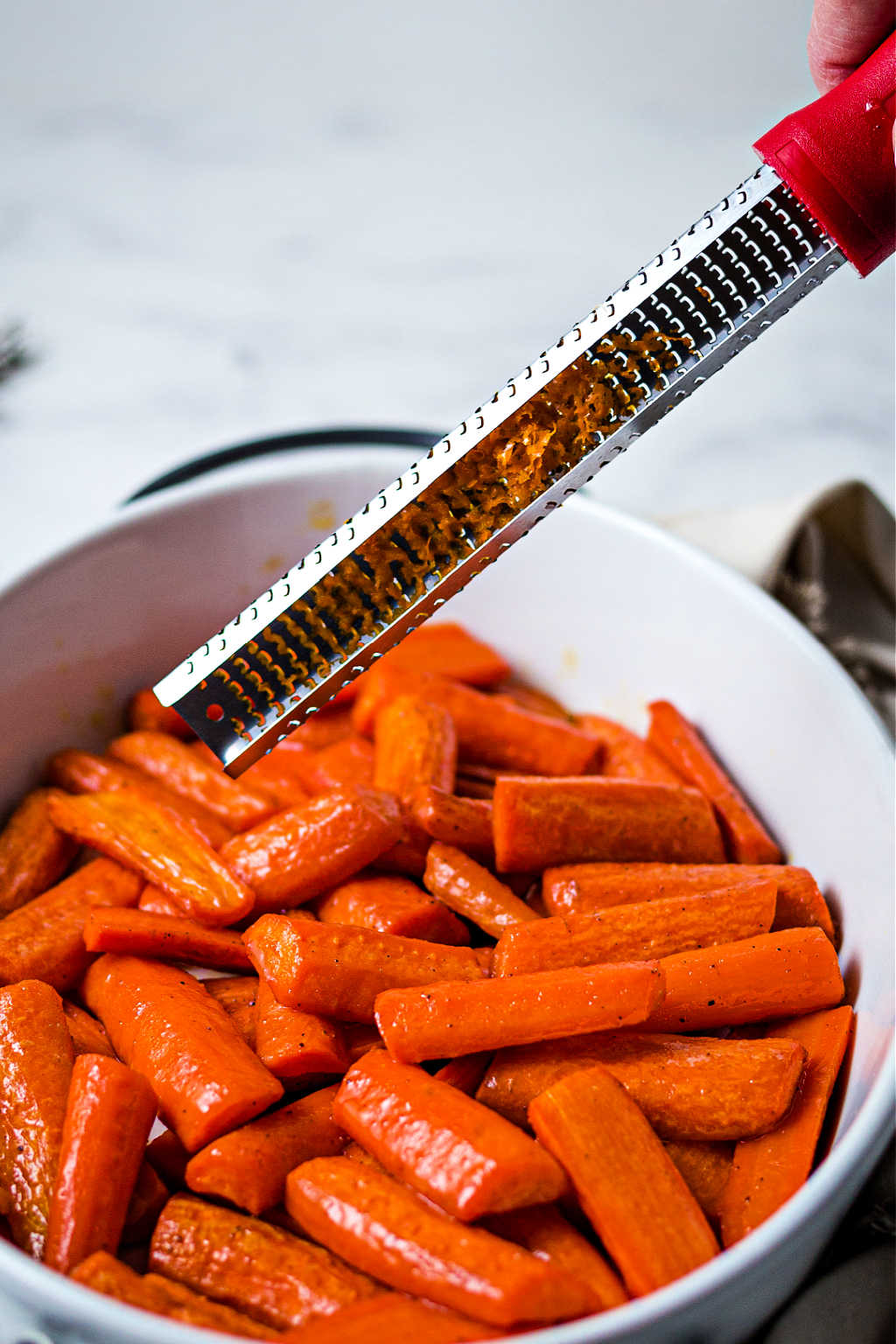 adding orange zest from a microplane zester to roasted carrots in a white dish.