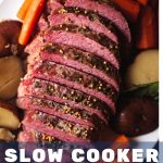 sliced slow cooker corned beef on a white platter with carrots and potatoes with a bowl of cabbage on a table.