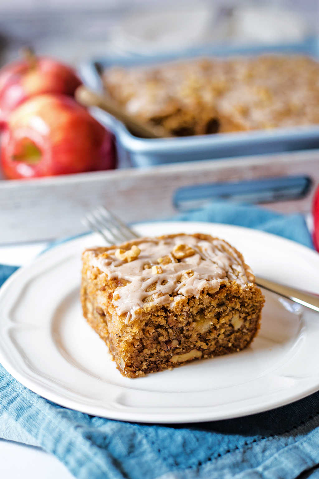 a slice of apple walnut cake on a white plate on a table with a tray of apples.