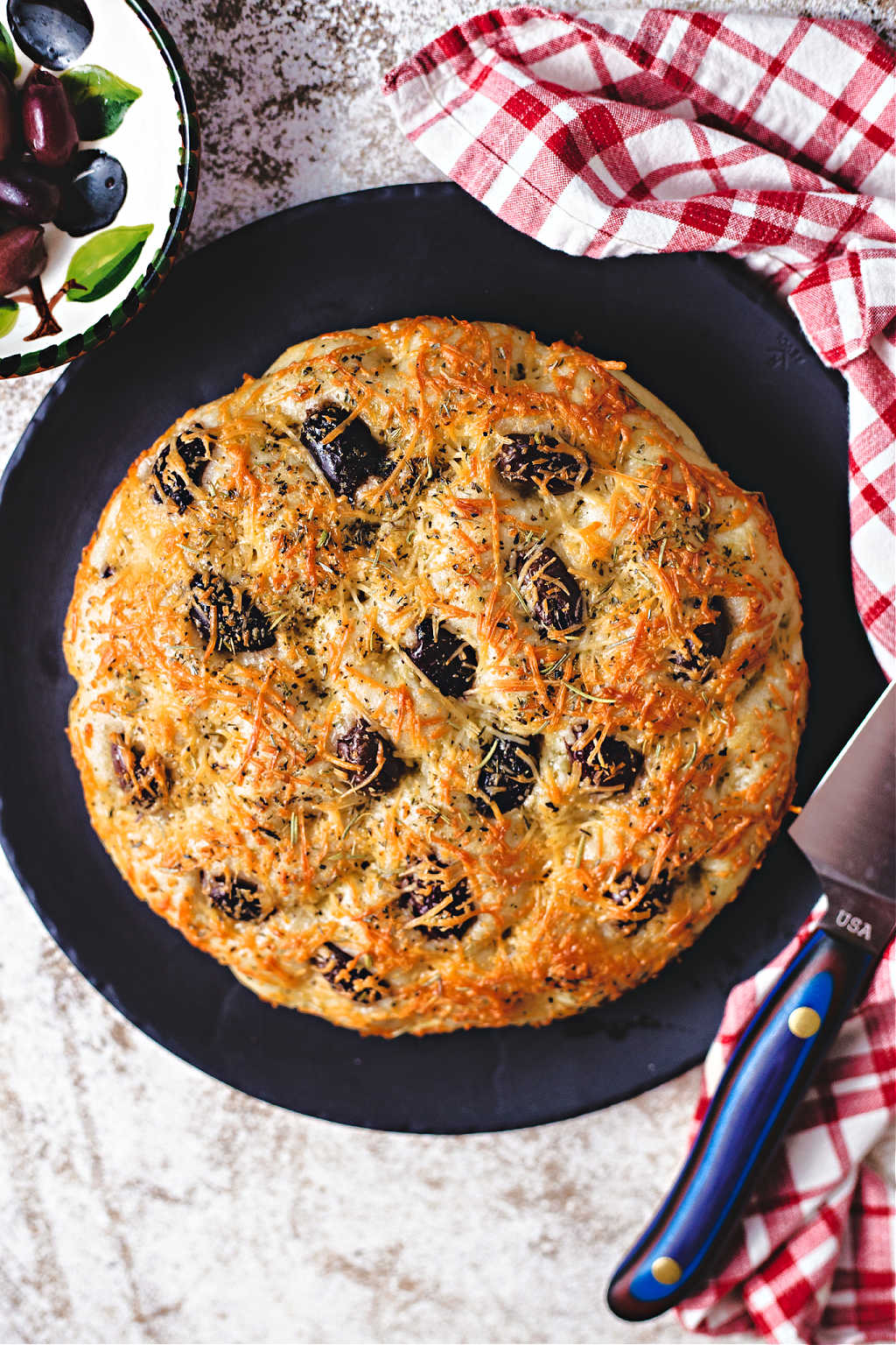 greek olive focaccia on a black stone platter with a carving knife.