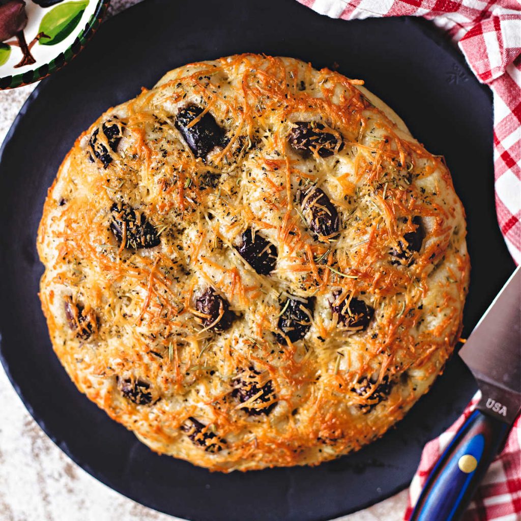 greek olive focaccia on a black stone platter with a carving knife.