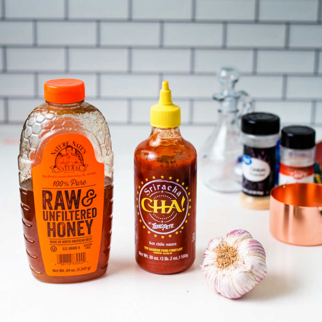 a bottle of honey, a bottle of sriracha, and a head of garlic on a kitchen counter.