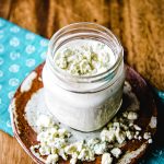 buttermilk blue cheese dressing in a mason jar with blue cheese crumbles on top sitting on a wooden table.