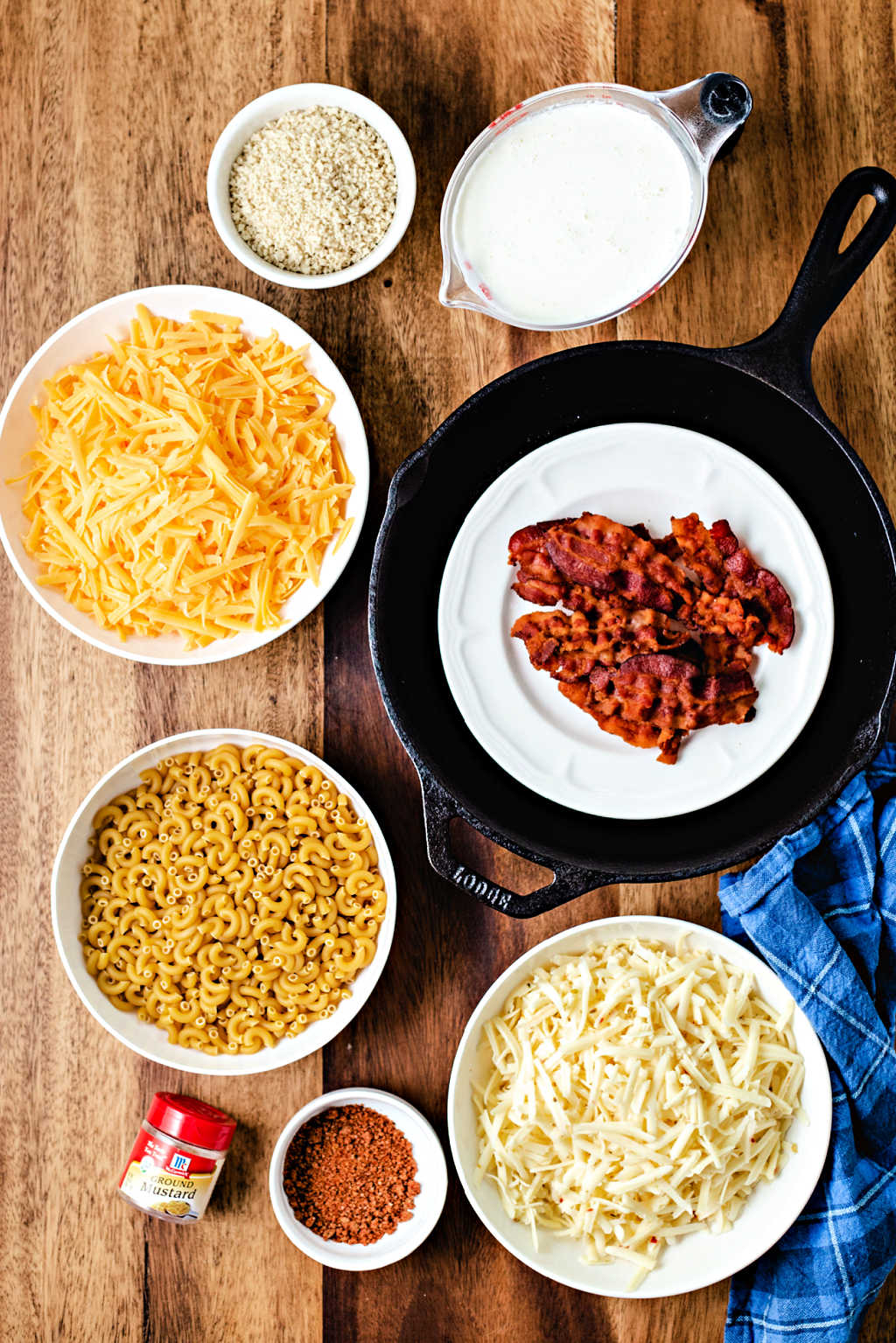 ingredients for smoked mac and cheese on a wooden table: pepper jack cheese, sharp cheddar cheese, bacon, dry macaroni, milk.