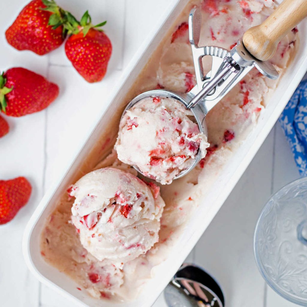 a container of homemade strawberry ice cream with an ice cream scoop and strawberries on a table.