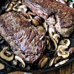 two New York strip steaks with mushrooms in a cast iron skillet.