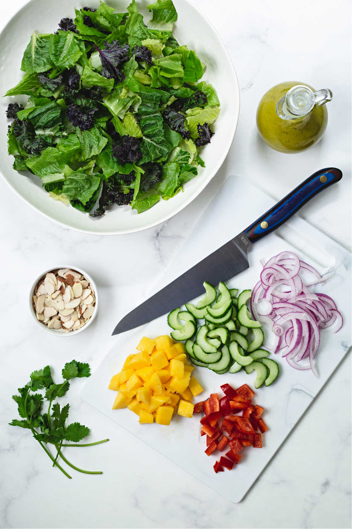 ingredients for grilled chicken salad on a counter: salad greens, sliced almonds, chopped onion, mango, red bell pepper, and cucumbers.