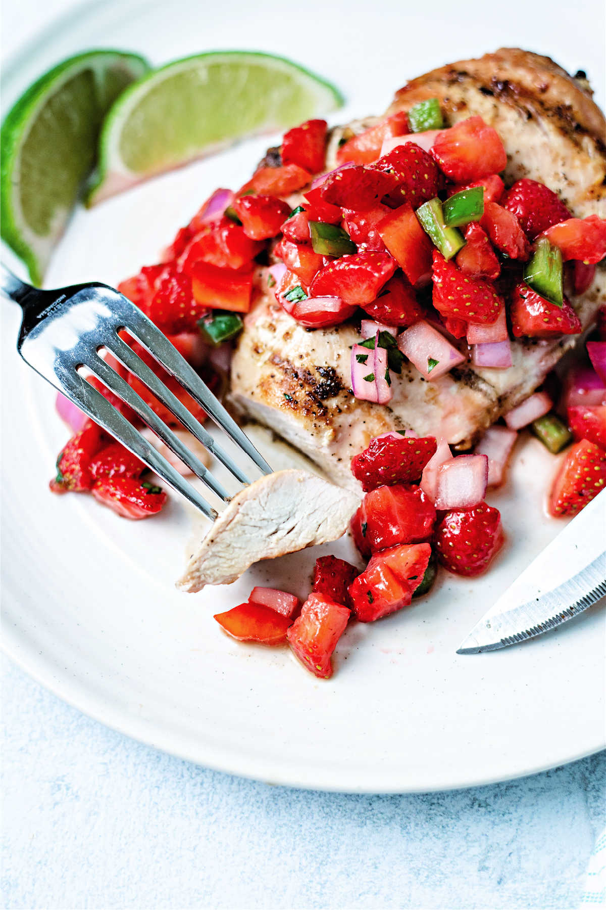 a fork with a piece of cut chicken breast beside the full serving on a white plate with strawberry salsa.