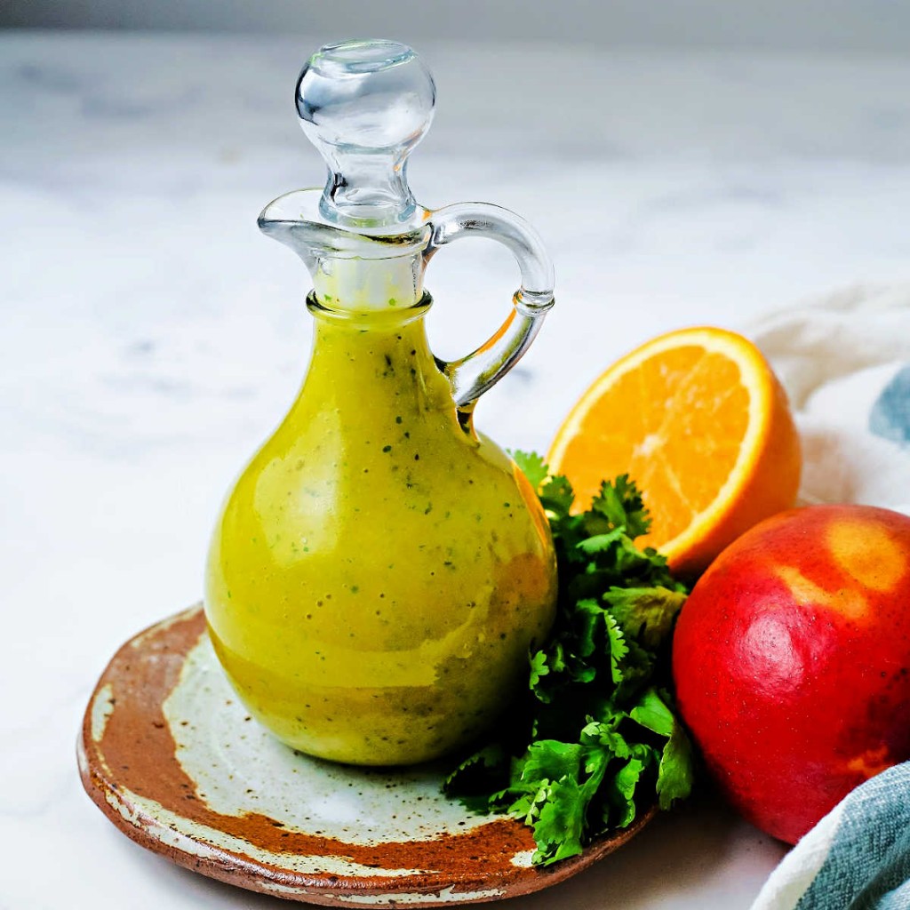 Adecorative carafe filled with mango dressing sitting on a table surrounded by cilantro, an orange slice, and a mango.